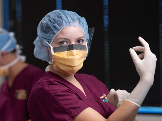 Young woman in mask, cap and maroon scrubs putting on gloves.