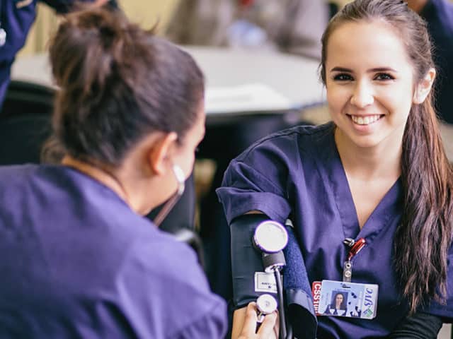 Two young women in scrubs using a blood pressure gauge.