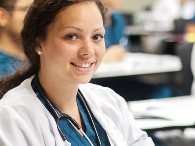Young woman in white smock over blue scrubs in classroom.